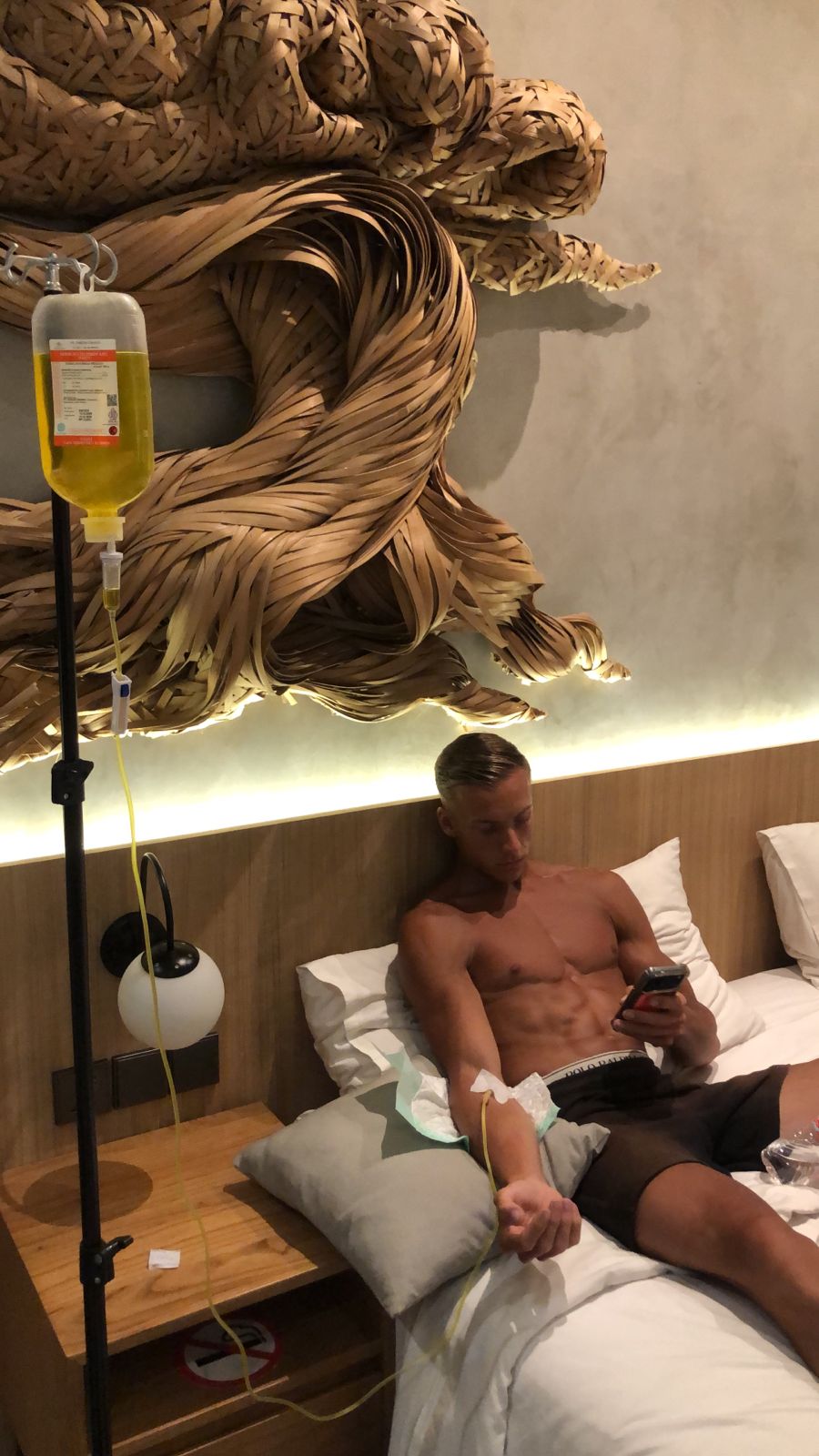 Shirtless man receiving a mobile IV drip therapy while relaxing in a hotel bed.